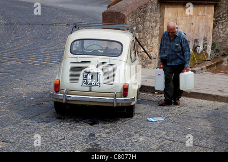 Altes Fiat 600 Auto und älterer Mann mit Tanks sammeln Wasser am Brunnen auf der Piazza Armerina, Sizilien, Italien. Sizilianischer Alltag in Sicilia, Italien Stockfoto