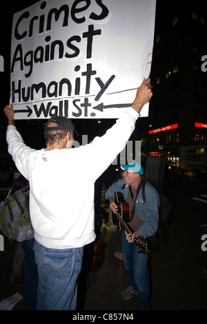 Mann hält ein Schild mit der Aufschrift "Verbrechen gegen die Menschlichkeit Wall Street", während Demonstranten bei Occupy Wall Street im Zuccotti Park Gitarrespielen Stockfoto