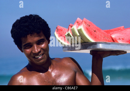 Rio De Janeiro, Brasilien. Lächelnd jungen Mann mit einem Tablett saftige Wassermelone Scheiben für Verkauf am Strand. Stockfoto