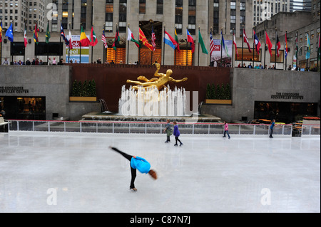 Weiblichen Schlittschuhläufer bewegen ein Bein vorne gold Prometheus Skulptur, Fahnen, "30 Rock", Rockefeller Center Ice Rink, New York, USA Stockfoto