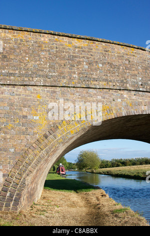 Narrowboat auf einem Kanal eingerahmt von Ziegelbrücke gewölbt und tiefblauen Himmel Hintergrund. Eine Sperre kann in Ferne zu sehen Stockfoto