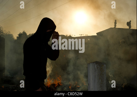 Dale Farm Räumung. Ein Demonstrant zu Fuß durch den Rauch von Bränden und Silhouette gegen frühen Morgensonne. Zwei Männer auf dem Dach. Stockfoto