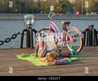 Eine Mädchen liegt auf einem hölzernen Damm von der Moskwa im Gorki Park an einem sonnigen Tag. In der Nähe befinden sich ihr Fahrrad und Spielzeug. Stockfoto