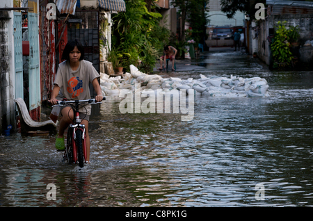 Thai Girl Fahrten mit dem Fahrrad durch Hochwasser, Sandsäcke im Hintergrund, der hochwasserschutzwand bei der überlaufende Phra Khanong Canal, Sukhumvit Road, Soi 50, Bangkok, Thailand am 30. Oktober 2011 zu verstoßen. Thailand erlebt die schlimmste Überschwemmung in mehr als 50 Jahren. Quelle: Kraig Lieb Stockfoto