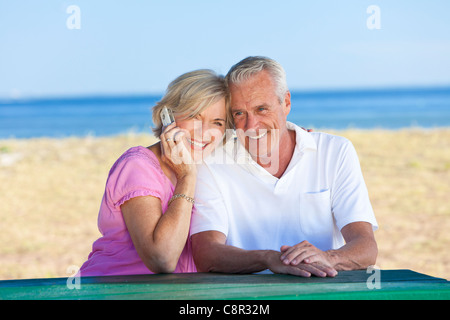 Ältere Mann und Frau Brautpaar sitzen zusammen an einem Tisch am Strand sprechen auf einem Handy Stockfoto