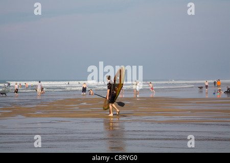 Bude Cornwall England Jüngling im Neoprenanzug tragen Kajak in Brandung bei Urlaubern, die in sanften Wellen im Meer spielen Stockfoto