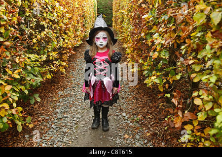 Kleines Mädchen mit Gesicht gemalt und in Hexenkostüm zu Halloween im Bild zwischen Hecken von Herbstlaub Stockfoto