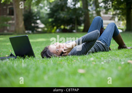 Niederlande, Maastricht, junge Frau liegt auf dem Rasen mit Laptop und Handy