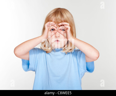 Junge mit langen blonden Haaren, die auf der Suche durch die Finger als Fernglas - isoliert auf grau Stockfoto