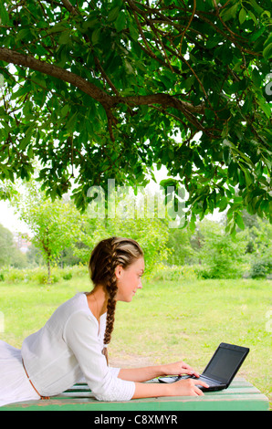 Frau ist ziemlich barfuß auf einer Bank unter dem grünen Baum legen. Sie arbeitet mit kleinen notebook Stockfoto