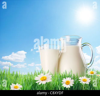 Milchkännchen und Glas auf dem Rasen mit Kamille. Auf einem Hintergrund von den sonnigen Himmel mit Wolken. Stockfoto