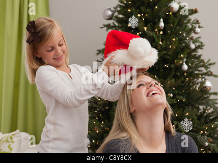Tochter (6-7) putting Weihnachtsmütze auf Mutters Kopf vor Weihnachtsbaum Stockfoto