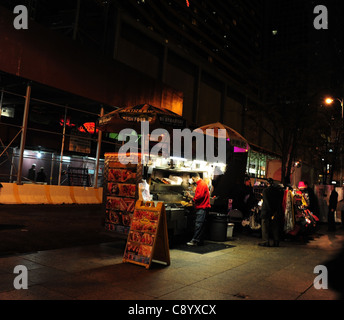 Nacht Schuss rote Jacke Mann Kochen Halal-Speisen bei einem Snack stand neben einem Verkauf Kleidung, West 52nd Street 7th Avenue, New York Stockfoto