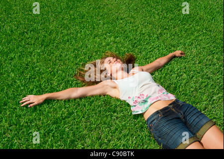 Junge Frau in kaukasischen 20er Jahre lange blonden Haaren liegen im grünen Rasen, Arme gestreckt, um ihre Seiten tragen Jeansshorts sorglos glücklich Stockfoto