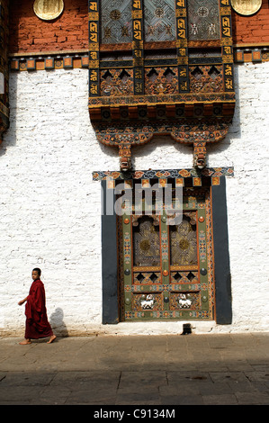 Ein junger Lehrling Mönch geht es vorbei an einigen herrlich geschnitzten und bemalten Fenstern in einem Innenhof der Punakha Dzong in Bhutan. Stockfoto