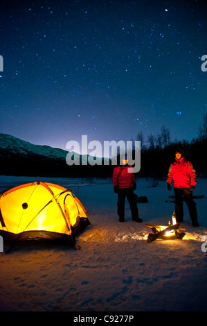 Zwei Männer wärmen sich am Lagerfeuer stehen neben ihrem beleuchteten Zelt, Eklutna Lake in Chugach State Park, Alaska Stockfoto