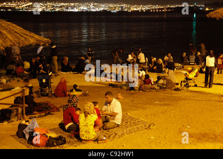 Menschen am öffentlichen Strand in der Nacht mit Eilat im Hintergrund, Aqaba, Jordanien. Stockfoto