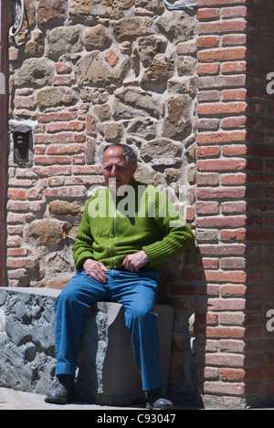 Eine italienische männliche Glatzenbildung Senioren sitzt auf einer steinernen Bank in der Sonne gegen einen Stein und Ziegel Wand in Panicale, Italien. Stockfoto