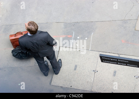 Ein Stadtarbeiter in einem Anzug steht mit Taschen auf der Straße und wartet unter Schildern für Moorgate und Liverpool Street Station in London, Großbritannien Stockfoto