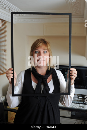 Eine Frau, die im Büro arbeitet und lächelnd durch einen leeren Bilderrahmen schaut Stockfoto