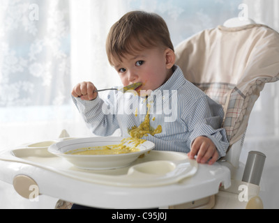 Ein und eine Hälfte Jahr alt junge in einem Hochstuhl sitzen und mit einem Löffel Suppe essen verschütten es auf seinem Hemd Stockfoto
