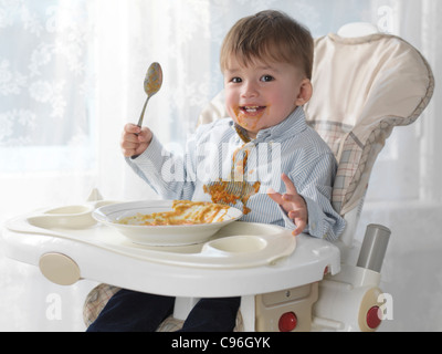 Ein und eine Hälfte Jahr alt fröhlicher Junge in einem Hochstuhl sitzen und mit einem Löffel Suppe essen verschütten es auf seinem Hemd Stockfoto