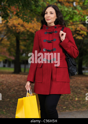 Lächelnde junge schöne Frau zu Fuß auf einem Bürgersteig trägt einen roten Mantel und hält Einkaufstaschen