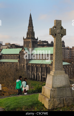 Ein Junge und ein Mädchen gehen vorbei an der keltischen Kreuz Grabstein in Glasgow Necropolis mit Glasgow Kathedrale im Hintergrund. Stockfoto