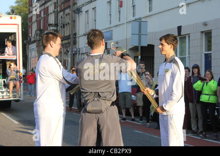 CARDIFF, UK, 26. Mai 2012. Ein Beamter überwacht wie Zak Lee Green (rechts) die Olympische Flamme an der nächsten Fackelträger übergibt. Dies geschieht über den "Kuss" wo sind die Fackeln zusammen gehalten, bis die Flamme die neue Fackel überträgt. (8. Tag des Fackellaufs) Stockfoto