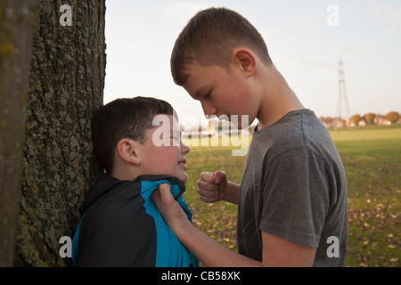 Ein älterer Junge Mobbing einen jüngeren Jungen mit seiner Faust holte hält ihn gegen einen Baum. Stockfoto