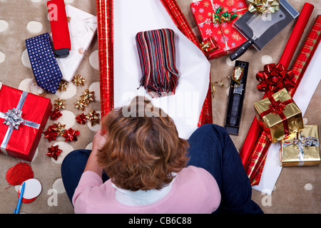 Obenliegende Foto einer Frau saß auf einem Teppich zu Hause verpacken ihre Weihnachtsgeschenke, Geschenke, Papier, Farbband und Bögen umgeben. Stockfoto