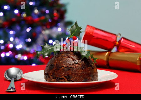 Foto von einer Christmas Pudding mit Holly an der Spitze mit Baum und Cracker im Hintergrund. Stockfoto