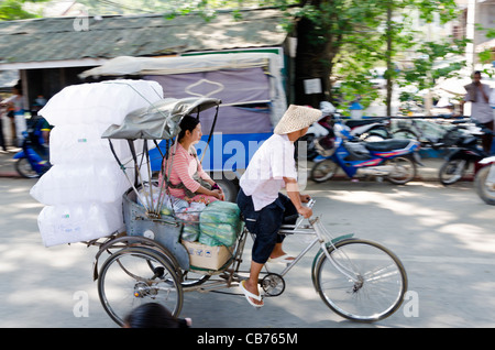 Ein Mann mit einem kegelförmigen Strohhut Pedale eine Velo-Rikscha mit großer Last & sitzende Frau im thailändisch-burmesischen Grenze in Tachileik Stockfoto