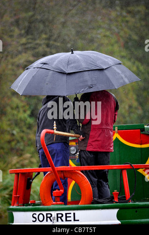 Zwei Narrowboater, die sich vor dem Regen am Steuer ihres Bootes auf dem Leeds Liverpool Canal, Yorkshire, Großbritannien, schützen. Stockfoto