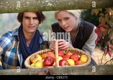 ein junges Paar posieren hinter eine hölzerne Barriere mit einem Flechtwerk Korb voller Äpfel Stockfoto