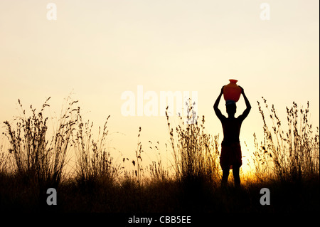 indischer Mann bei Sonnenuntergang mit einem Wassertopf auf diesem Kopf in der indischen Landschaft wandern. Silhouette Stockfoto