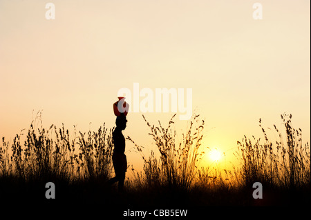 indischer Mann bei Sonnenuntergang mit einem Wassertopf auf diesem Kopf in der indischen Landschaft wandern. Silhouette Stockfoto