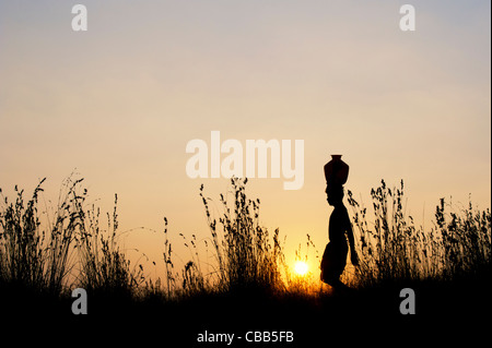 indischer Mann bei Sonnenuntergang mit einem Wassertopf auf diesem Kopf in der indischen Landschaft wandern. Silhouette Stockfoto