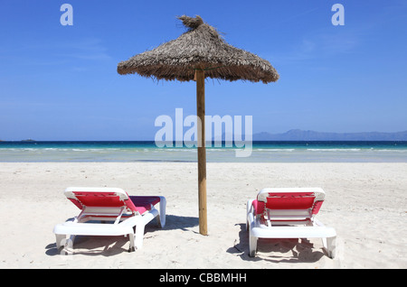 Zwei rosa gepolsterten Sonnenliegen und ein Stroh Sonnenschirm an einem breiten Sandstrand. Aufnahme in Alcudia, Mallorca, Spanien Stockfoto