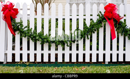 Garland und rote Schleifen hängen von einem weißen Lattenzaun für die Weihnachtszeit. Stockfoto