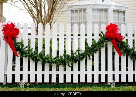 Garland und rote Schleifen hängen von einem weißen Lattenzaun für die Weihnachtszeit. Stockfoto