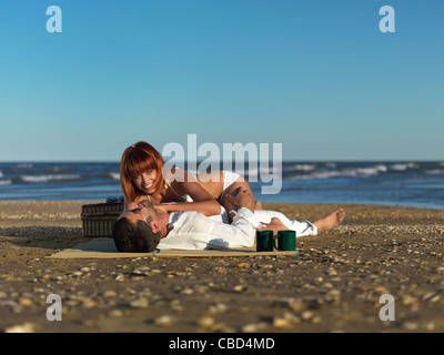 glückliches, junges Paar, in weiß gekleidet, entspannend, mit einem Picknick am Meeresufer Stockfoto