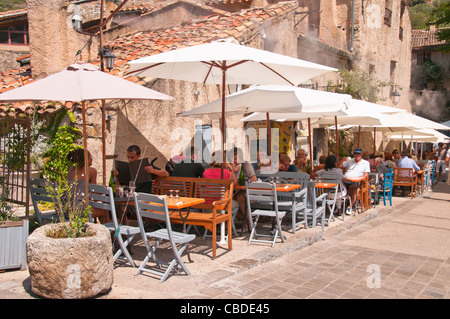 Holidaymakes unter Sonnenschirmen sitzen und genießen Sie Getränke in der Hill Top Dorf von St. Guilhem le Desert, Hérault, Languedoc Stockfoto