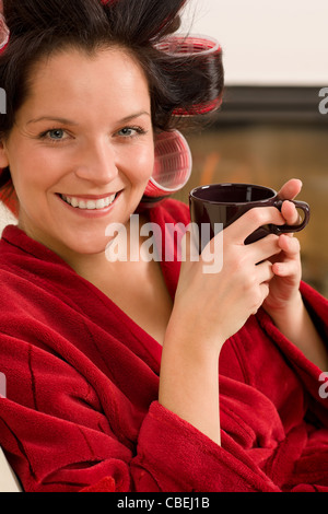Frau mit Lockenwickler halten Becher sitzen Kamin roten Bademantel Stockfoto