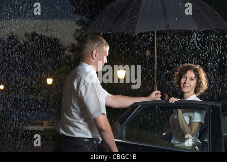 Ein Mann, eine Autotür öffnen und einen Regenschirm über eine Frau zu halten Stockfoto