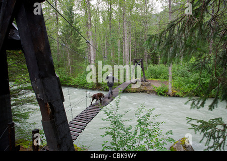 Mensch und Hund eine Hängebrücke über den Taiya River überqueren. Canyon City (Geisterstadt). Chilkoot Trail. Alaska. USA Stockfoto