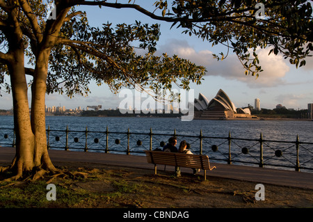 Ein paar sitzt auf einer Parkbank, den frühen Abend Blick auf Circular Quay und dem Opernhaus von Milsons Point Stockfoto