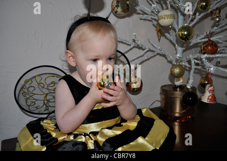 Babymädchen saugen Weihnachtsbaum Weihnachtskugel gekleidet in einem Bumble Bee Kostüm passenden Baum. Christbaumkugel-baby Stockfoto