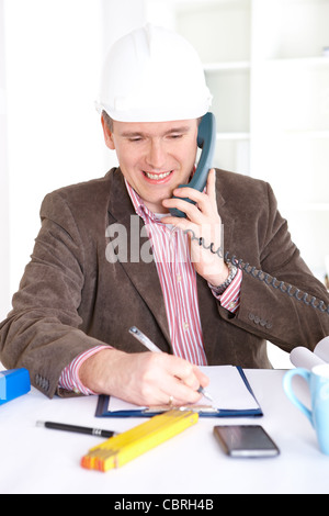 Architekt auf der Suche im Büro am Schreibtisch arbeiten. Tragen Helm und unter Notizen auf Papier mit einem Telefon in der hand. Stockfoto