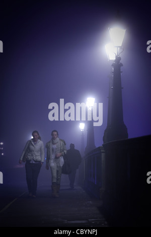 Zwei Frauen, bald gefolgt von einem Mann Fuß über Kingston Bridge in Richtung Hampton Wick an einem nebligen Abend. Stockfoto